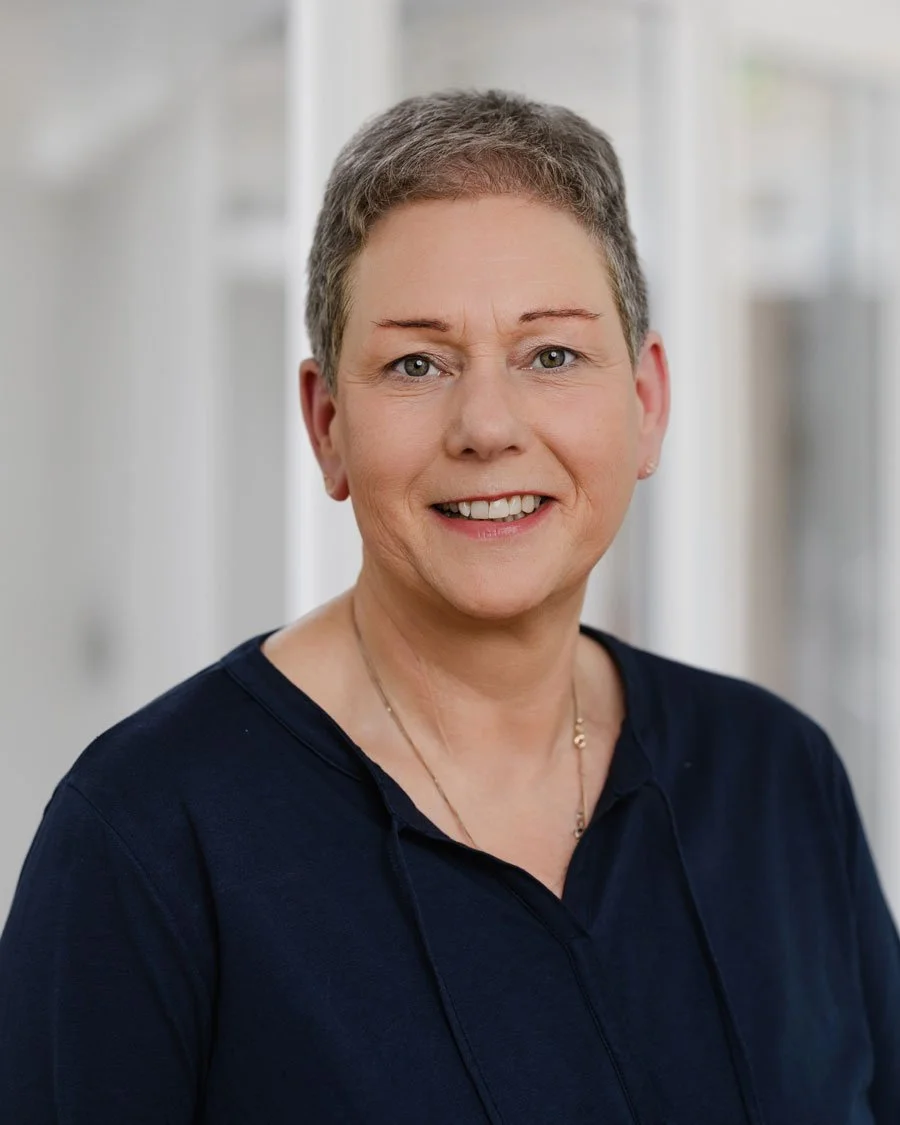 Nahaufnahme einer lächelnden, älteren Frau mit kurzen, grauen Haaren, trägt dunkle Kleidung und eine Halskette, vor unscharfem hellen Hintergrund.