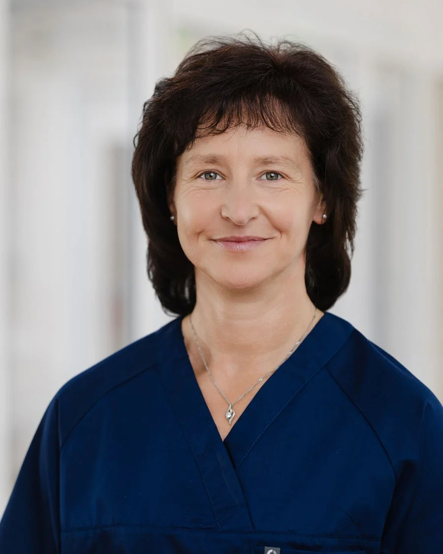 Porträt einer Frau in blauer Uniform, lächelnd, mit dunklem, kurz gestyltem Haar und einer silbernen Halskette