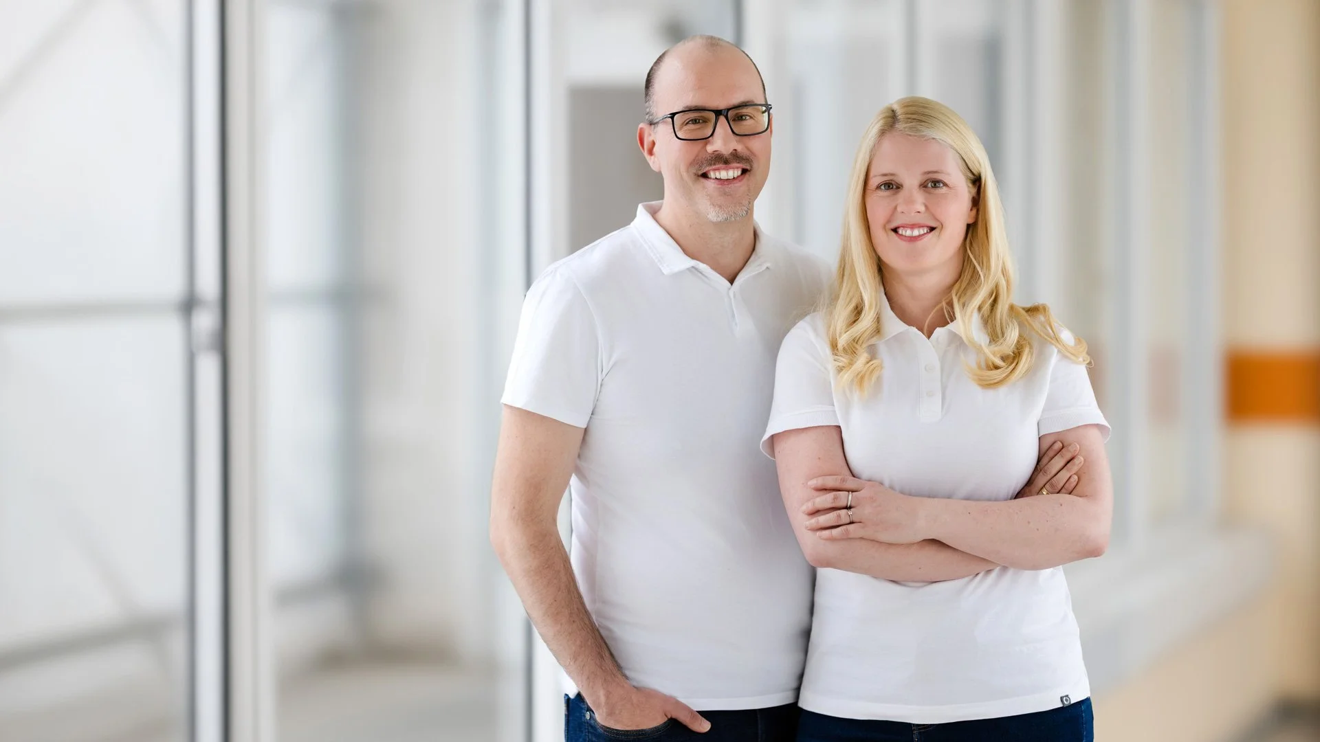 Zwei leicht lächelnde Menschen, ein Mann und eine Frau in weißen Polohemden, stehen in einem hellen, modernen Büroflur. Der Mann trägt Brille und hat kurze dunkle Haare, die Frau hat lange blonde Haare. Sie stehen vertrauensvoll nebeneinander, die Arme verschränkt.