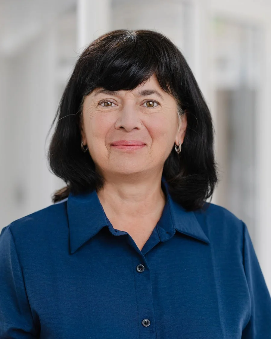 Nahaufnahme einer Frau mit schwarzen Haaren, die eine blaue Bluse trägt, vor einem neutralen Hintergrund.