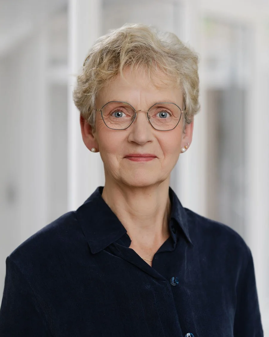 Porträt einer älteren Frau mit kurzen, blonden Haaren, Brille und Ohrringen, die eine dunkle Bluse trägt, vor einem hellen Hintergrund.
