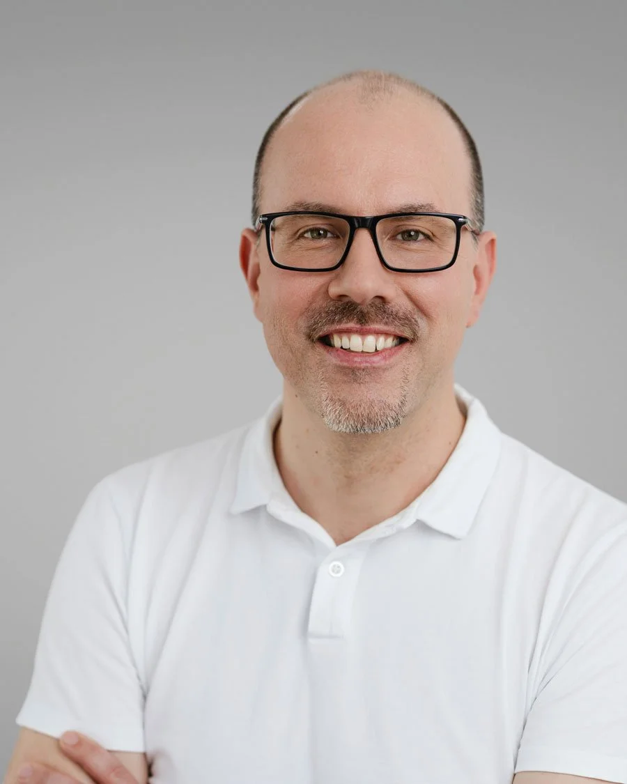 Porträt eines lächelnden Mannes mit Brille in einem weißen Hemd vor hellem Hintergrund.