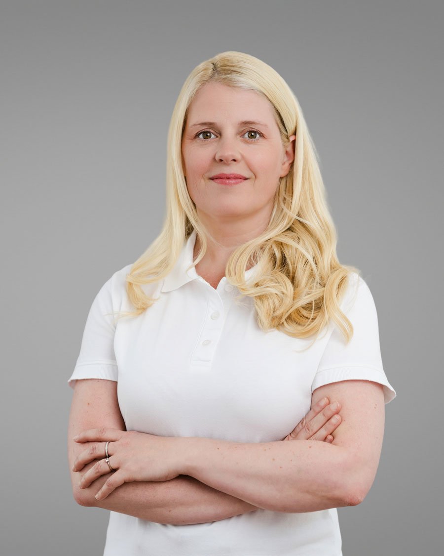 Junge blonde Frau mit lockigem Haar trägt ein weißes Polo-Shirt, steht vor grauem Hintergrund, mit verschränkten Armen, lächelt leicht.