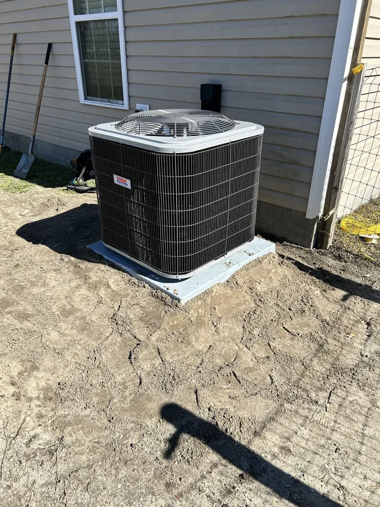 An outdoor air conditioning unit installed next to the side of a house with beige siding, a window, and some tools leaning against the house.