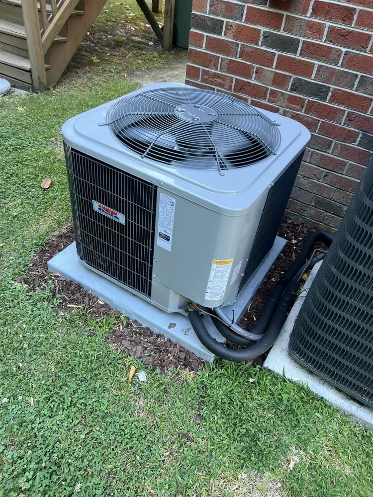 Outdoor air conditioning unit installed next to a brick wall on a concrete pad, with grass and mulch surrounding it.