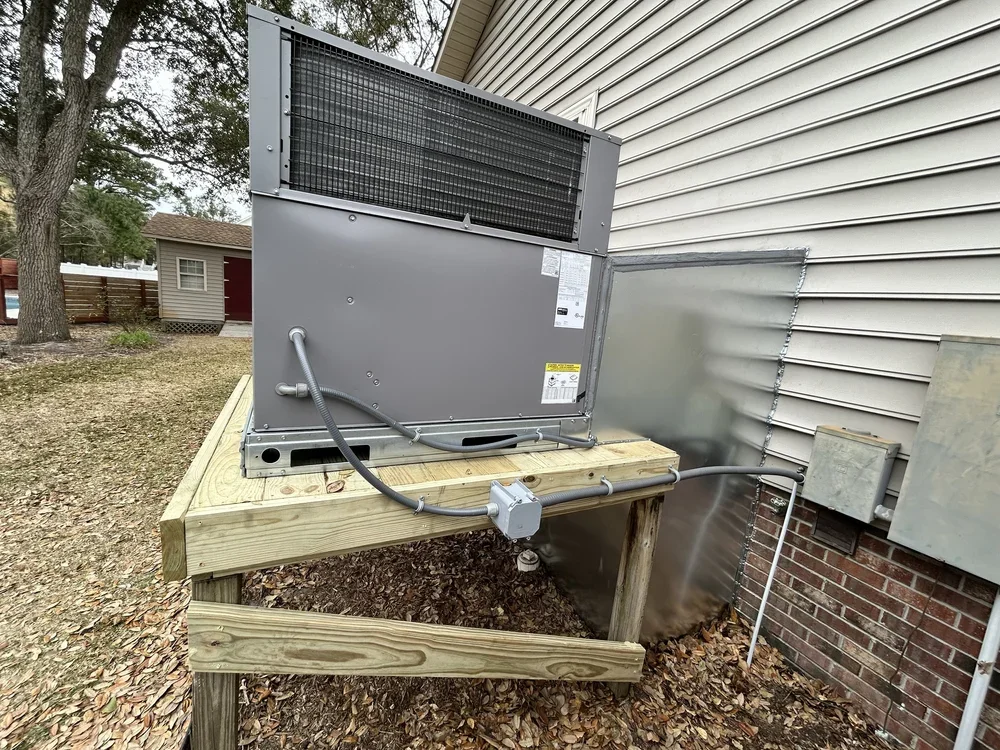 An outdoor HVAC unit installed on a wooden platform next to a house with vinyl siding and an electrical box.