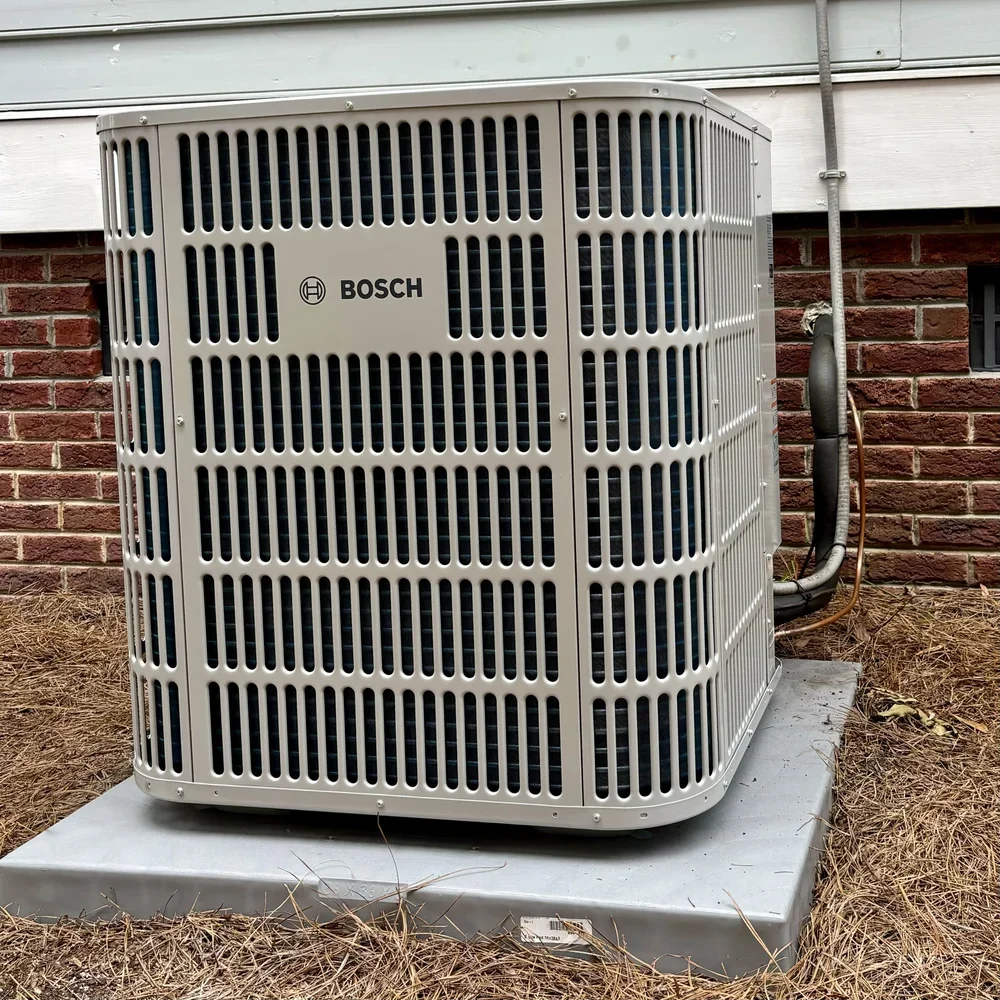 Outdoor Bosch central air conditioning unit on a concrete pad next to a brick wall.