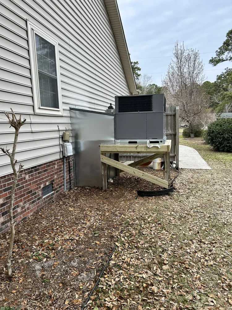 Backyard scene with house siding, a small leafless tree, and an air conditioning unit on a wooden platform with gravel and fallen leaves on the ground, a pathway, fence, and trees in the background.