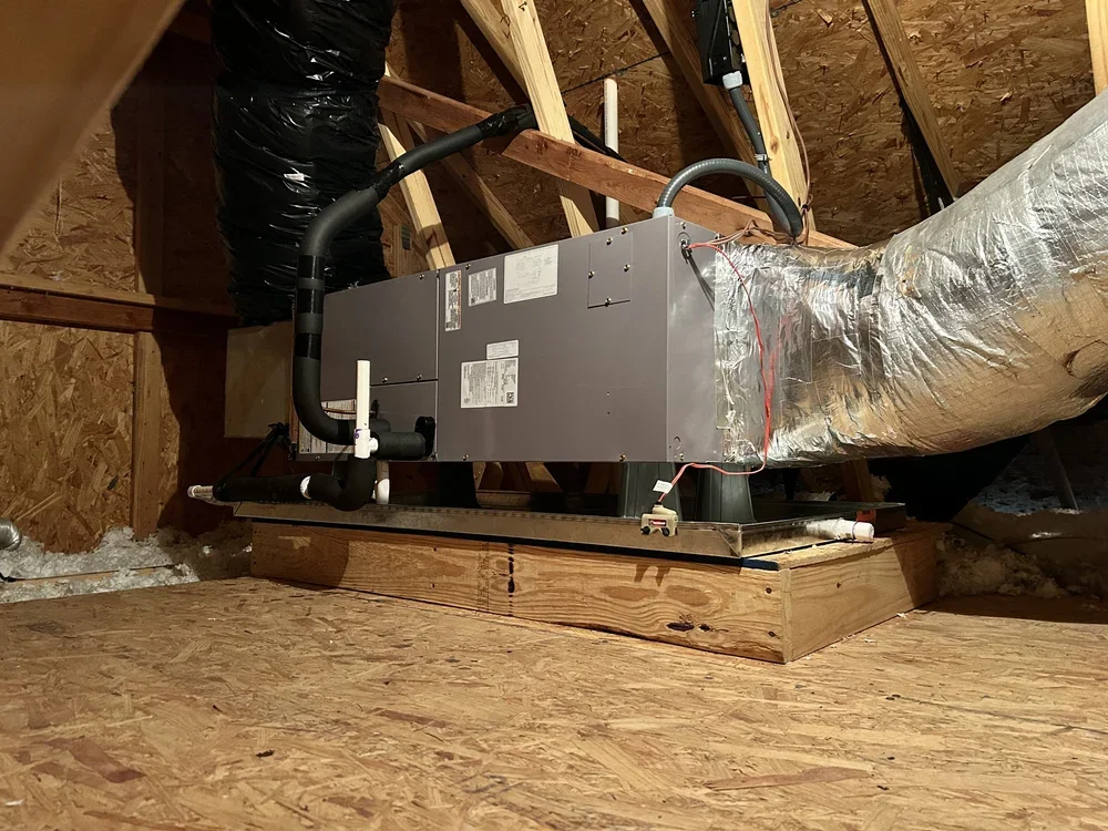 An HVAC air handler unit installed on an attic floor, with ductwork and insulation visible in the background.