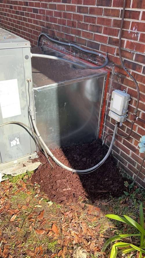 A metallic outdoor HVAC unit near a brick wall, with electrical connections and cables, and freshly dug soil at its base.