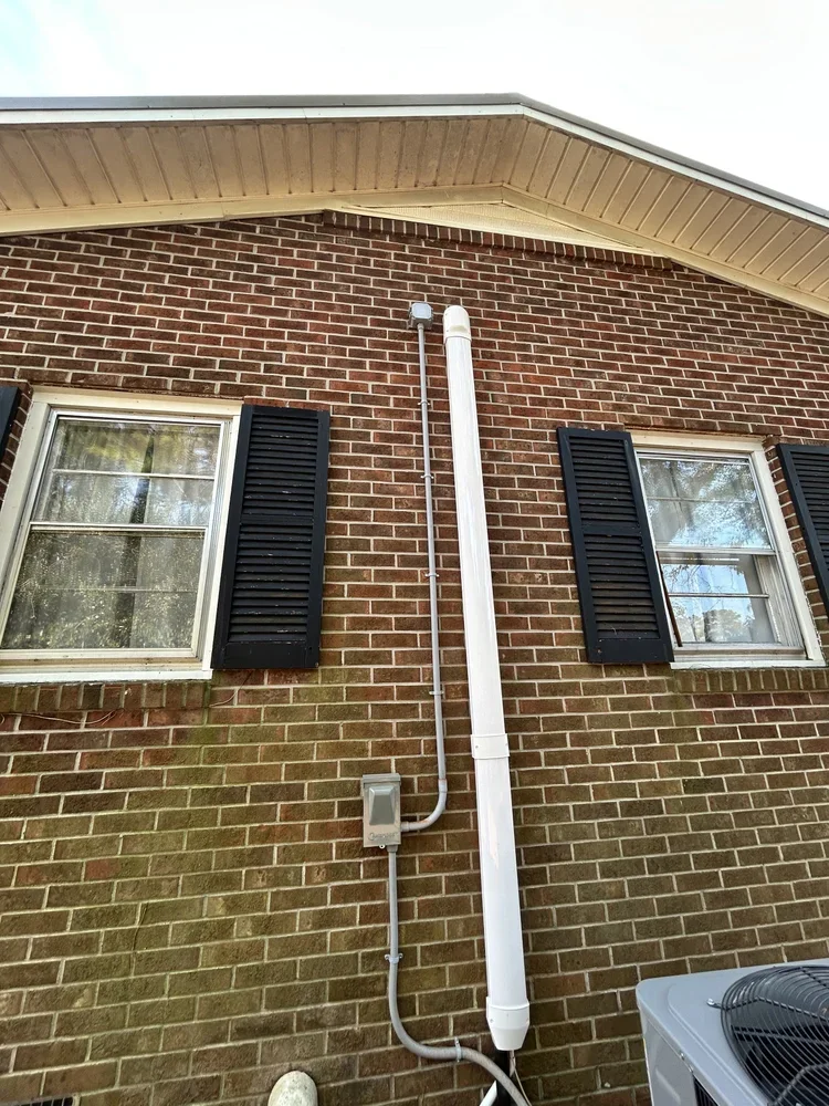 Exterior brick wall of a house with two windows and black shutters, and a white downspout attached to the wall. An electrical box and conduit are also visible. Part of an air conditioning unit is in the lower right corner.