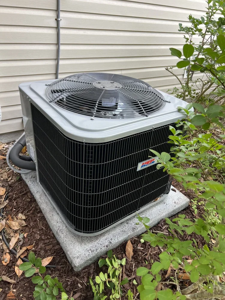 An outdoor HVAC air conditioning unit installed on a concrete pad beside a house with beige siding and surrounded by green bushes.