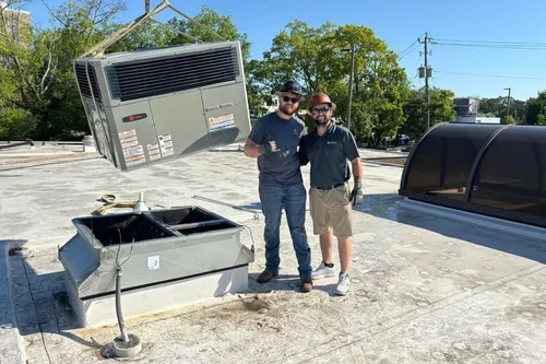 Two technicians standing on a rooftop next to an HVAC unit, with one unit removed and hanging in the air, and the other unit on the roof. The background features trees, power lines, and clear sky.