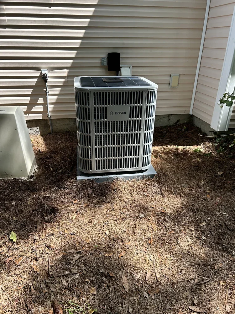 Outdoor view of a Bosch air conditioning unit installed next to the house wall, on a concrete slab sidewalk, with dirt and pine straw on the ground.