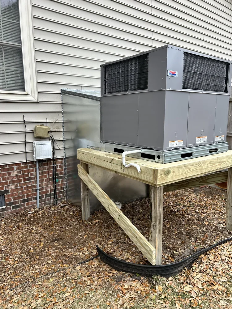 An outdoor HVAC unit on a wooden platform beside a house with beige siding and a window.
