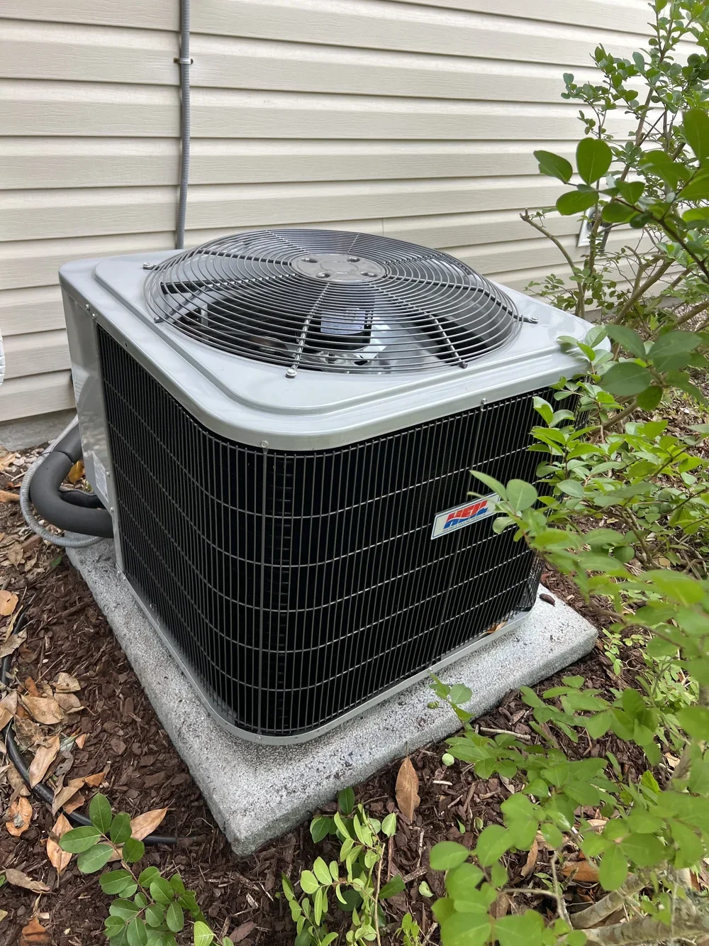 An outdoor air conditioning condenser unit with a protective grille on top, installed next to a beige vinyl siding house with some green bushes and brown mulch around it.