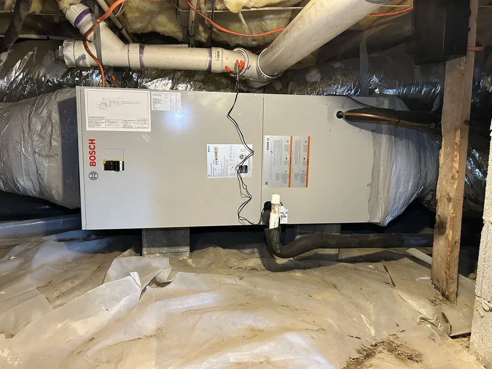 A Bosch furnace installed in a basement with exposed pipes and insulation, sitting on a concrete floor with plastic covering.