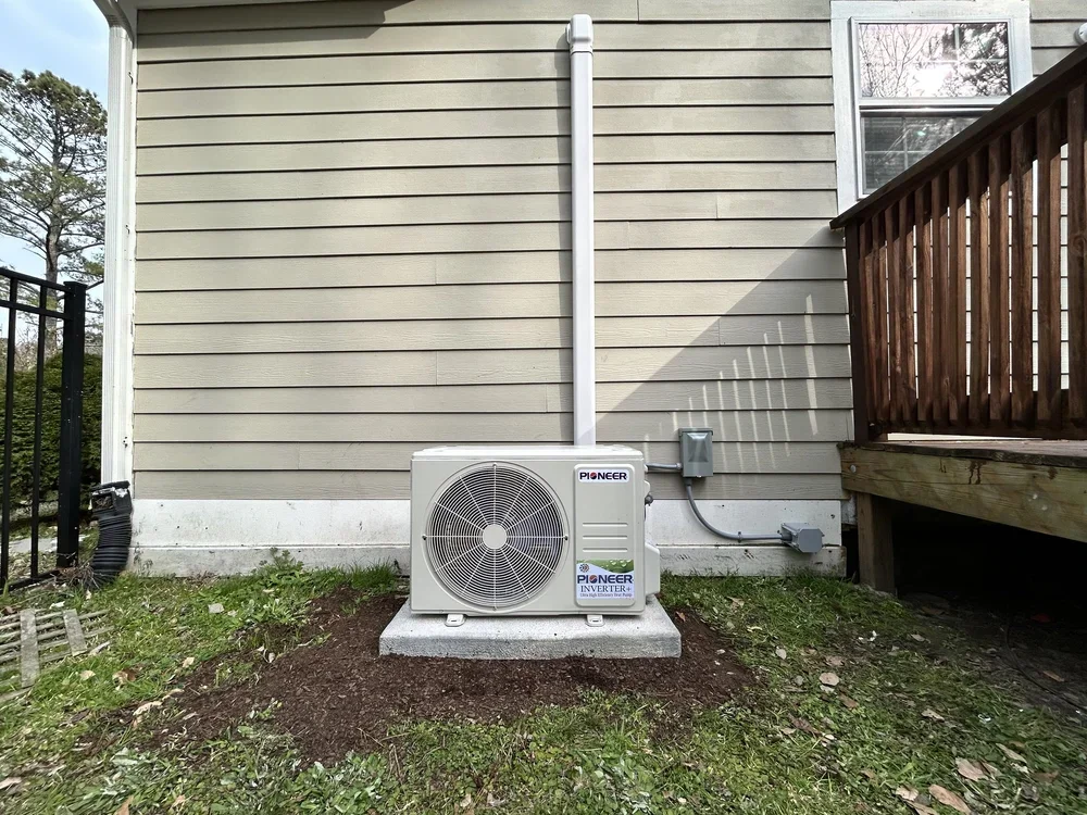 Exterior of a house showing an HVAC unit on a concrete pad, with electrical wiring and conduit, a small window, and part of a wooden deck with railing.