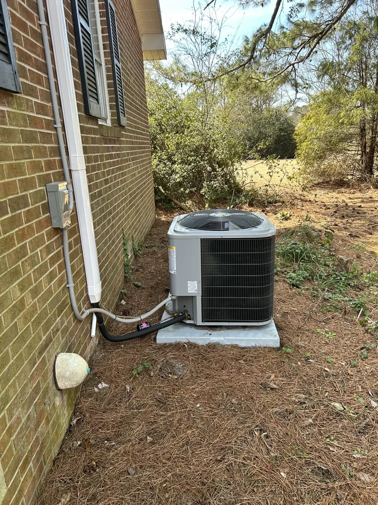 An outdoor air conditioning unit installed next to a brick house on a concrete pad with trees and bushes in the background.