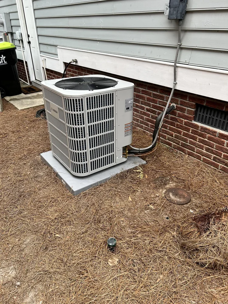 An air conditioning unit installed outside a house on a concrete pad, with a house wall, door, and some outdoor items in the background.