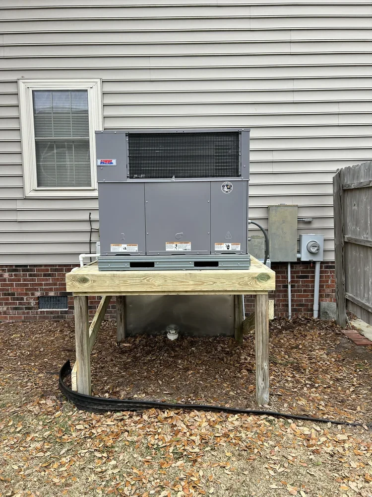 An outdoor HVAC unit on a wooden platform next to a house wall, with fallen leaves on the ground.