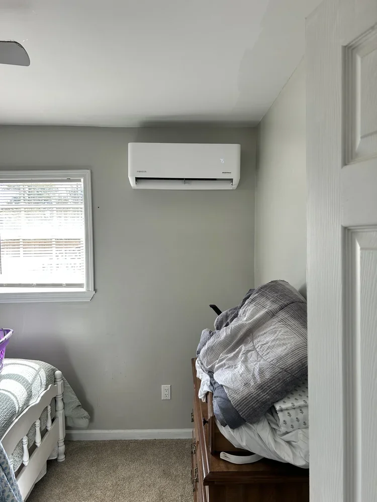 A bedroom with a window, an air conditioner mounted high on the wall, and a wooden chest with a pile of folded bedding and a remote control on top.