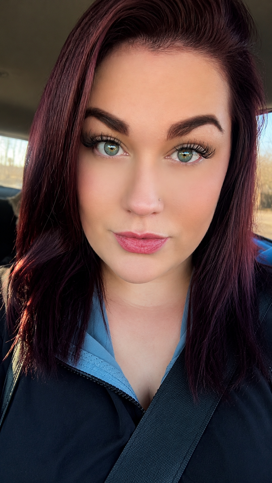 A woman with long, maroon hair and blue eyes. She has makeup, including eyeliner and lipstick, and wears a black jacket with a blue sports top underneath. She is seated in a vehicle, wearing a seatbelt, with a blurred outdoor background.