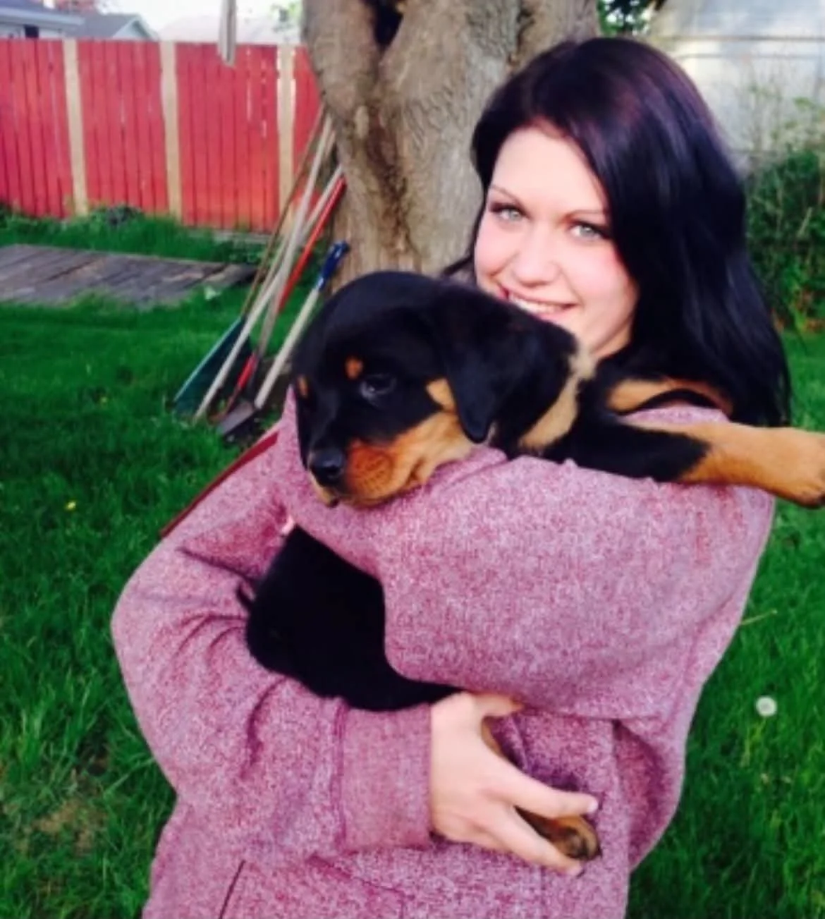 A woman with black hair smiling and holding a Rottweiler puppy in a backyard with a tree, grass, and a red fence in the background.