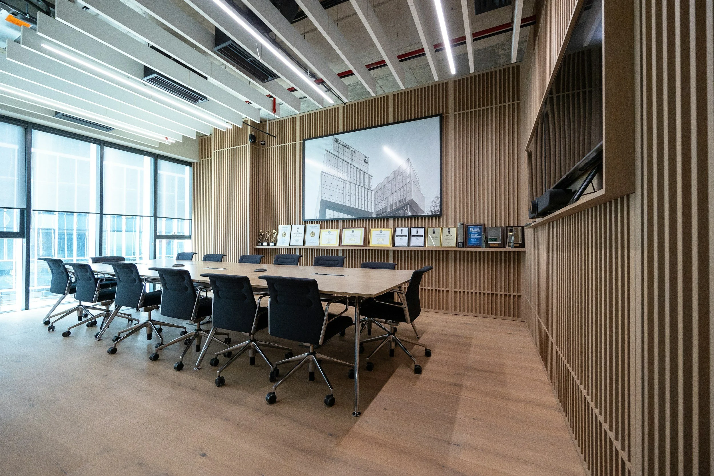 Modern conference room with a large wooden table surrounded by black office chairs, a wood-paneled wall with framed certificates or awards, a big screen displaying building architecture plans, and large windows allowing natural light to enter.
