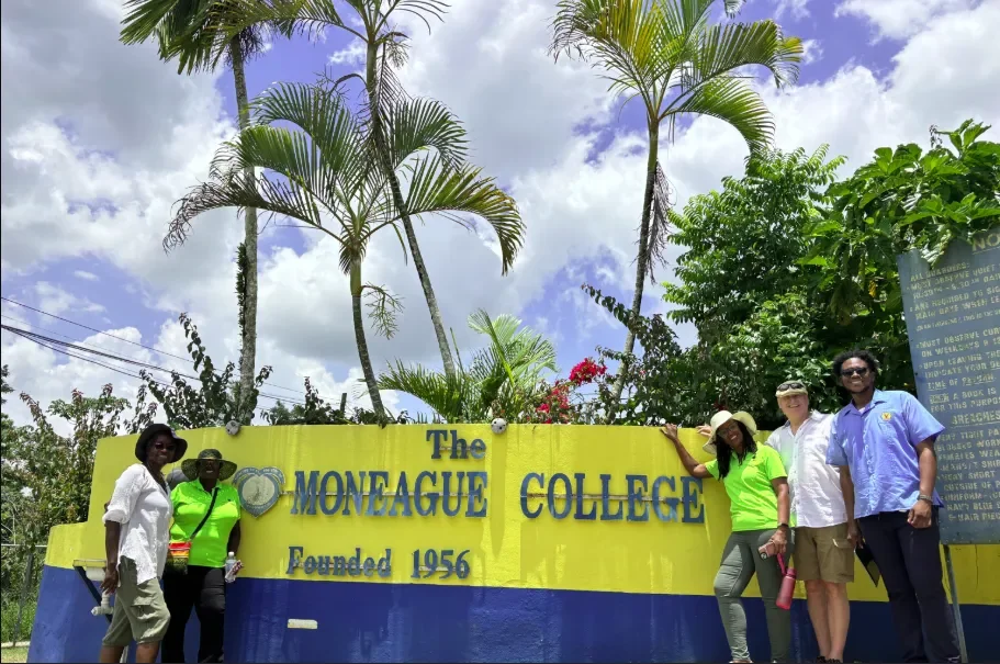  Jamaica Culinary Tours group and local guides posing in front of the bright yellow entry sign at The Moneague College in St. Ann during the Roots &amp; Routes cultural heritage tour. 