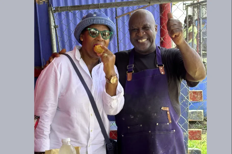  Jamaica Culinary Tours guest enjoying a hot fried dumpling while posing with a smiling local street food chef giving a thumbs-up on the Roots &amp; Routes tour. 