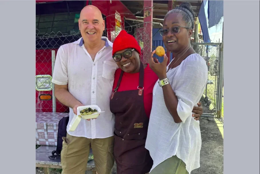  Jamaica Culinary Tours guests smiling with a local street food vendor while holding fresh Jamaican callaloo and a hot fried dumpling on the Roots &amp; Routes tour. 