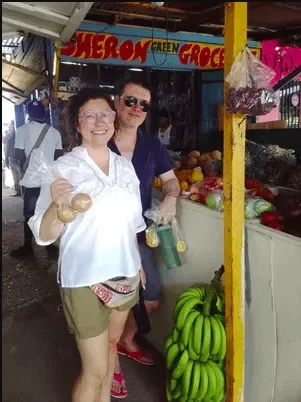 A tourist couple smiling while holding fresh local produce outside "Sheron's Green Grocery" market stall on the Roots & Routes food tour in Jamaica.