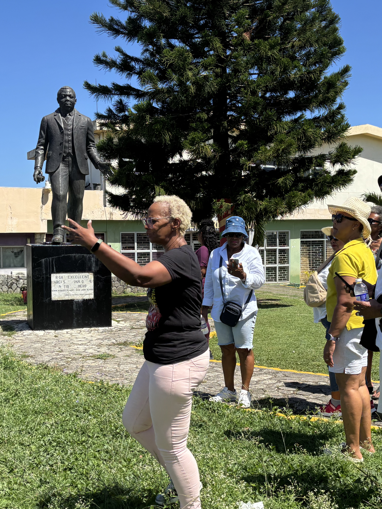 Tour guide gesturing toward a bronze statue of Marcus Garvey while a group of visitors listens nearby.