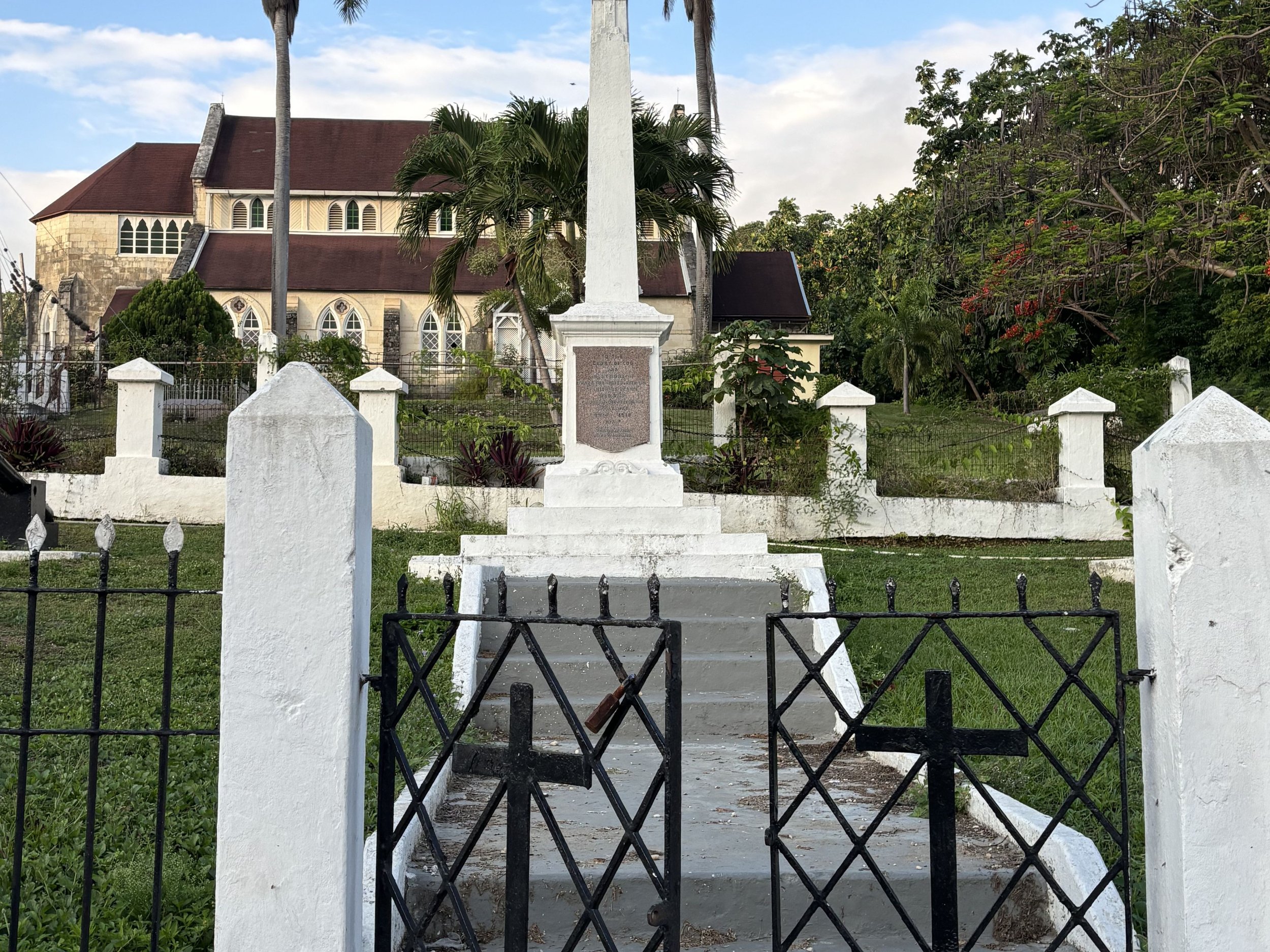 White stone monument surrounded by an iron gate, with a historic church building in the background.
