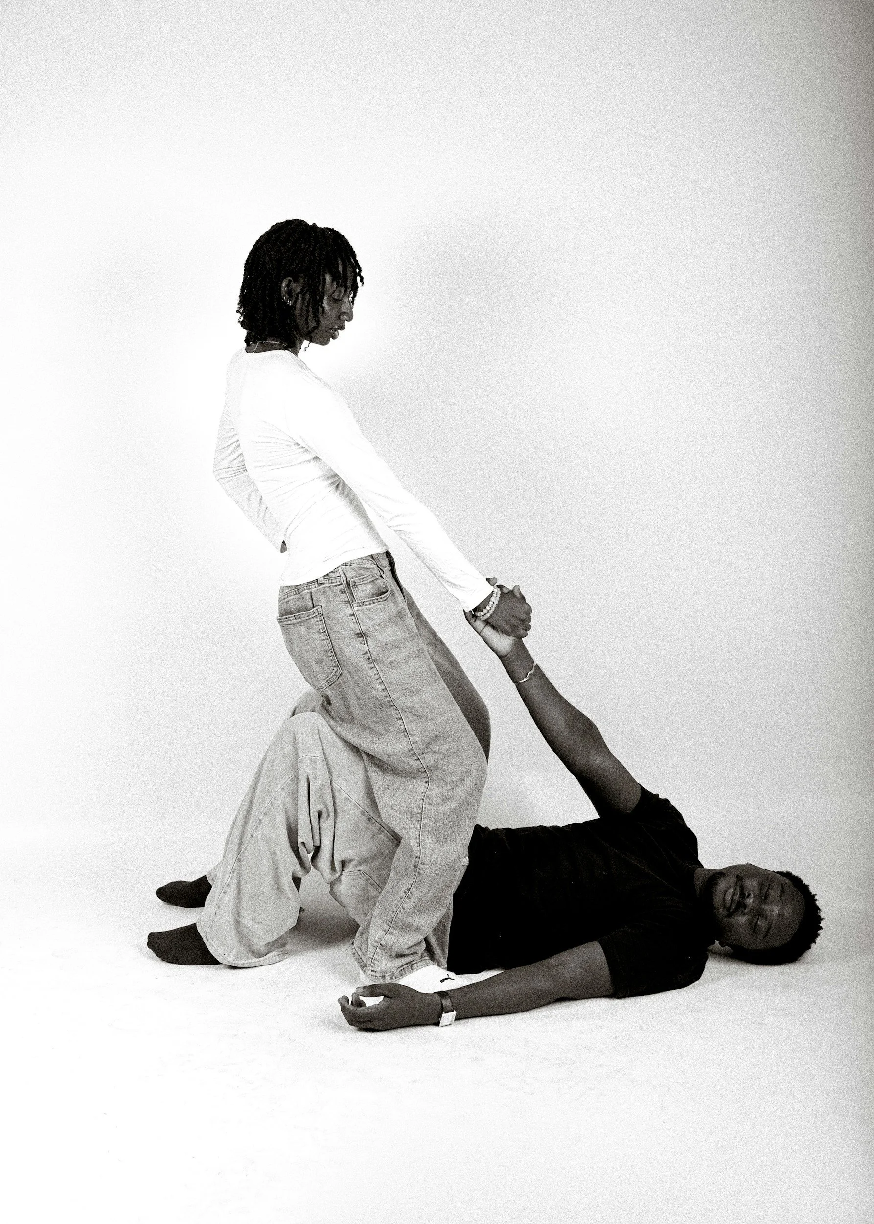 Black and white photo of four people engaged in a collaborative pose, with one person lying on the ground and two others standing and supporting them, against a plain background.