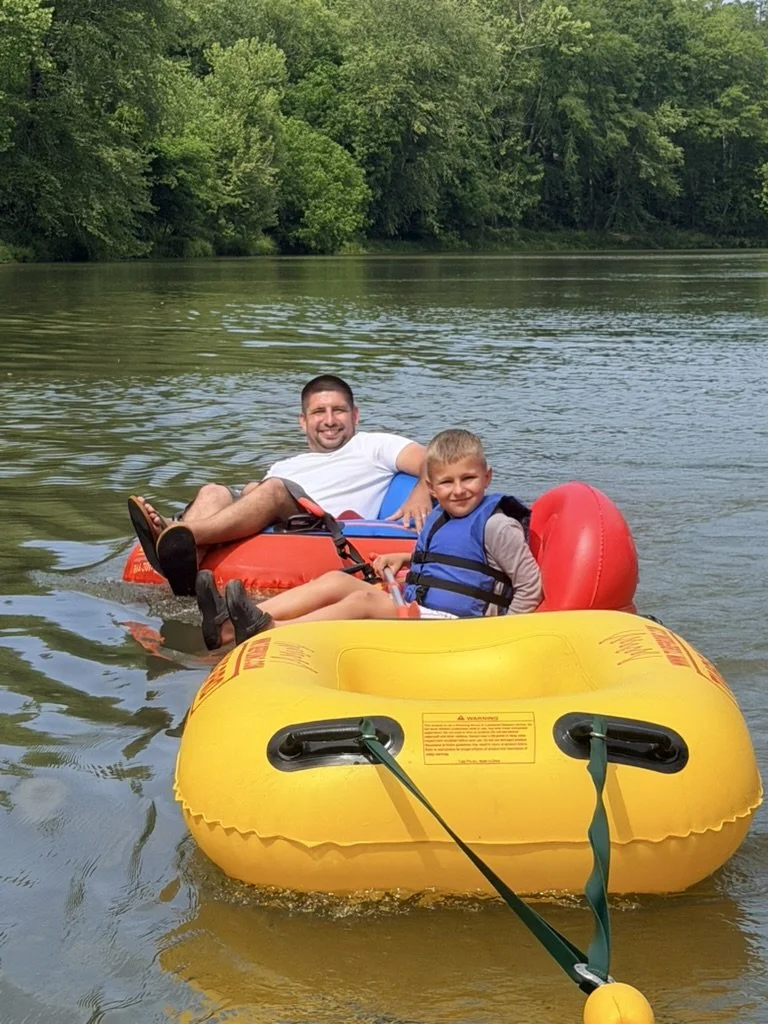 A man and a young boy floating on a river in an orange inflatable tube and a small red boat, surrounded by green trees.