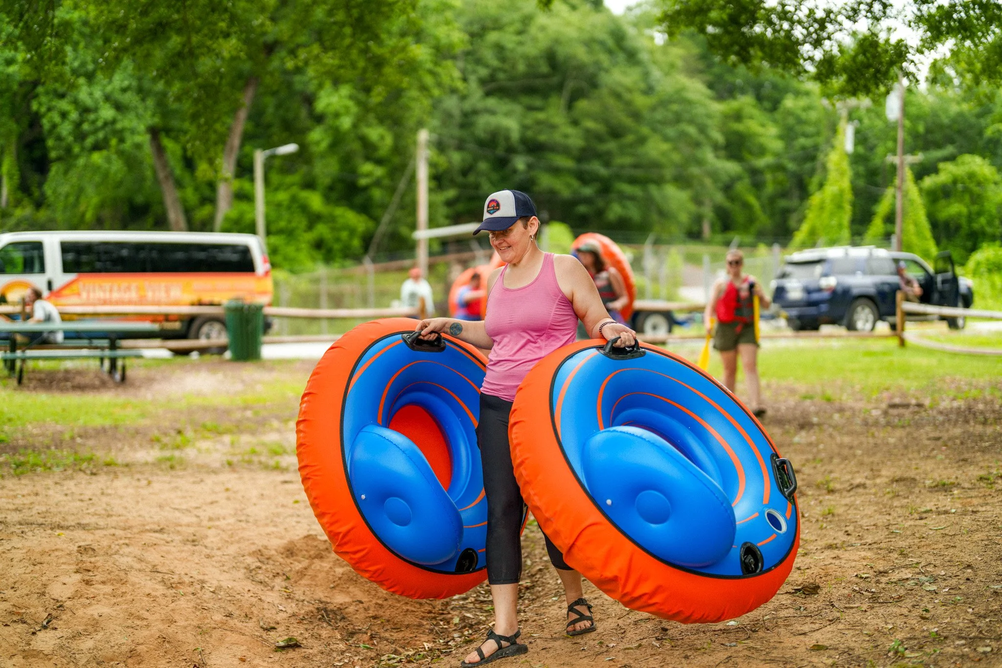 Woman in pink tank top and black pants carrying two large blue and orange inflatable tubes outdoors on a dirt path with trees and people in the background.