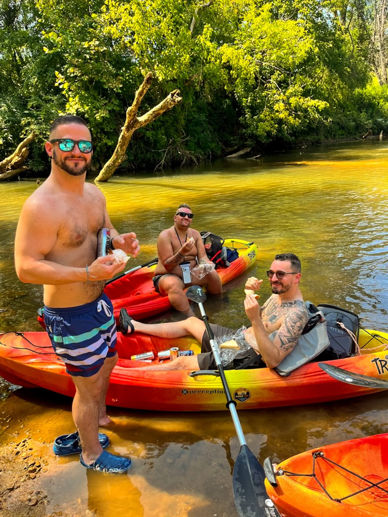 Four men enjoying a day kayaking on a river surrounded by lush green trees, with two of them eating snacks in the water, one standing in wet shoes and the others sitting in kayaks.