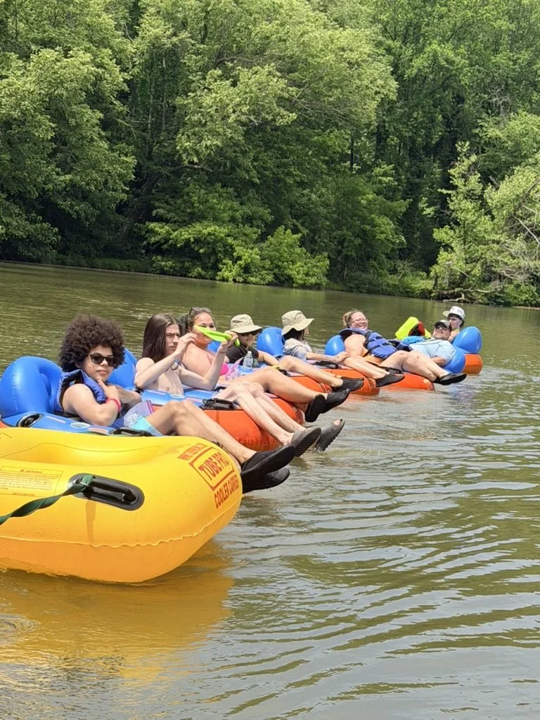 Group of people floating on the river in colorful inner tubes, surrounded by lush green trees.