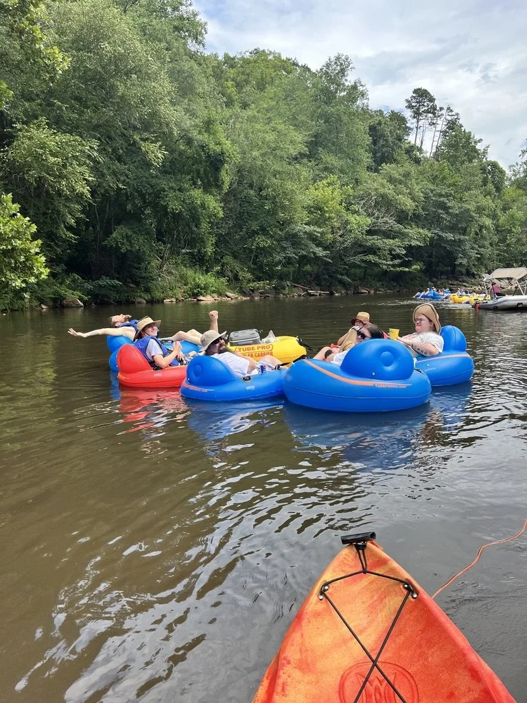 A group of five people in colorful inflatable tubes floating on a river, wearing hats and sunglasses, with lush green trees in the background. The photo is taken from a kayak at the river's edge.