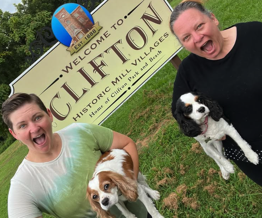 Two women smiling and holding Cavalier King Charles Spaniel dogs in front of a sign that says 'Welcome to Cliffton Historic Mill Village, Home of Clifton Park and Beach' on a grassy area.