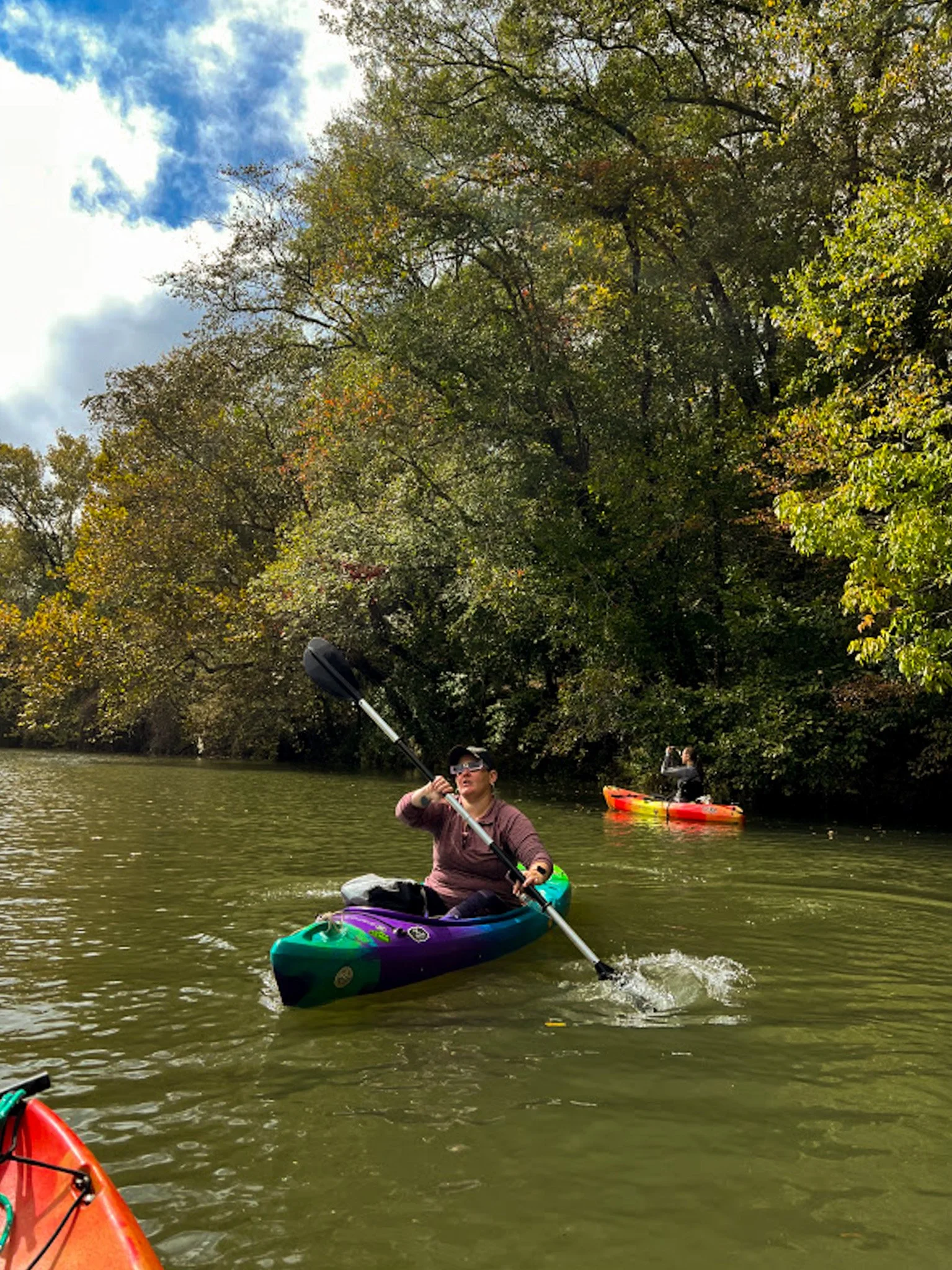 Person kayaking on a river surrounded by trees with green and yellow leaves under a partly cloudy sky.