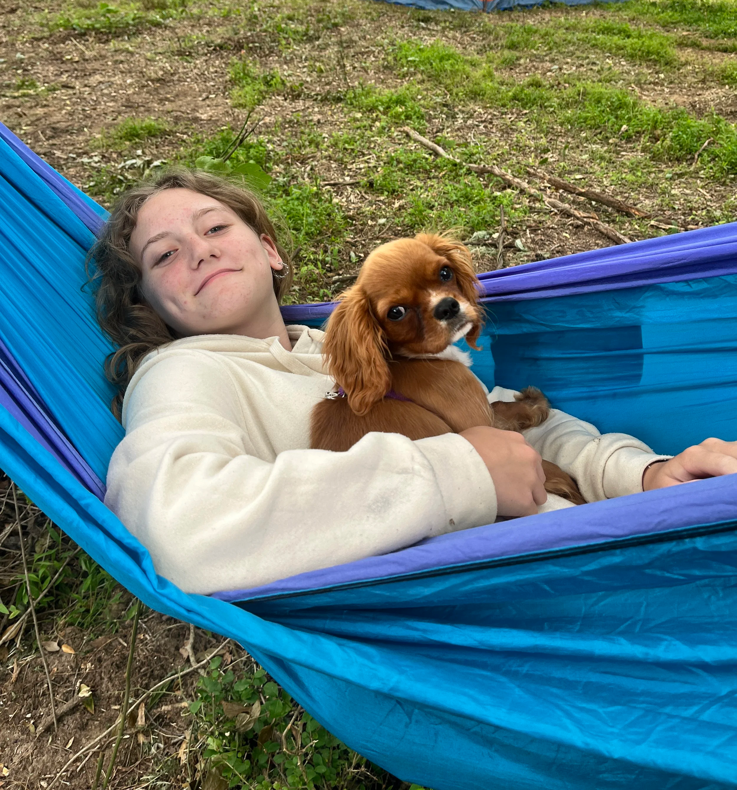A young girl lying in a blue hammock outdoors with a small brown and white dog resting on her chest. The background shows soil and green plants.