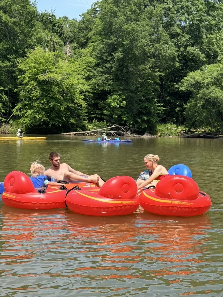 Family enjoying relaxing on red and blue inflatable boat in a calm river with green trees in the background.