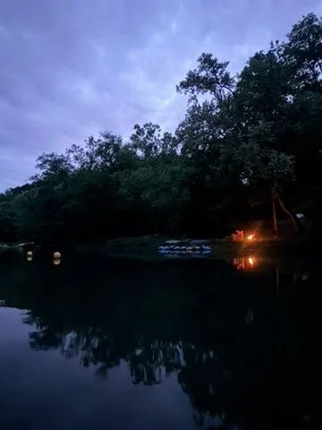 Calm river at dusk with trees on the bank and a small campfire glowing in the distance.