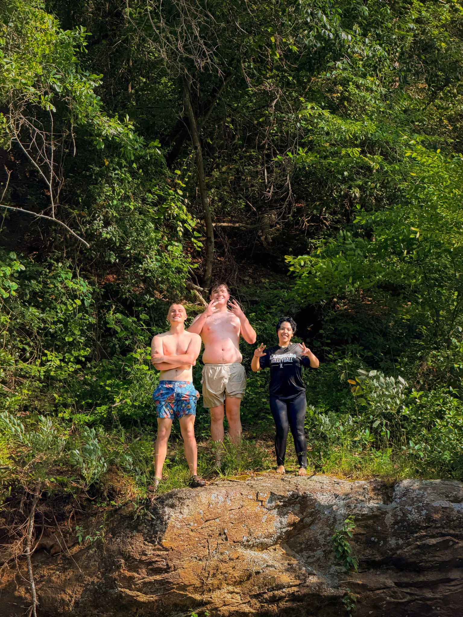 Three people standing on a rock ledge in front of dense greenery and trees, smiling and making gestures.