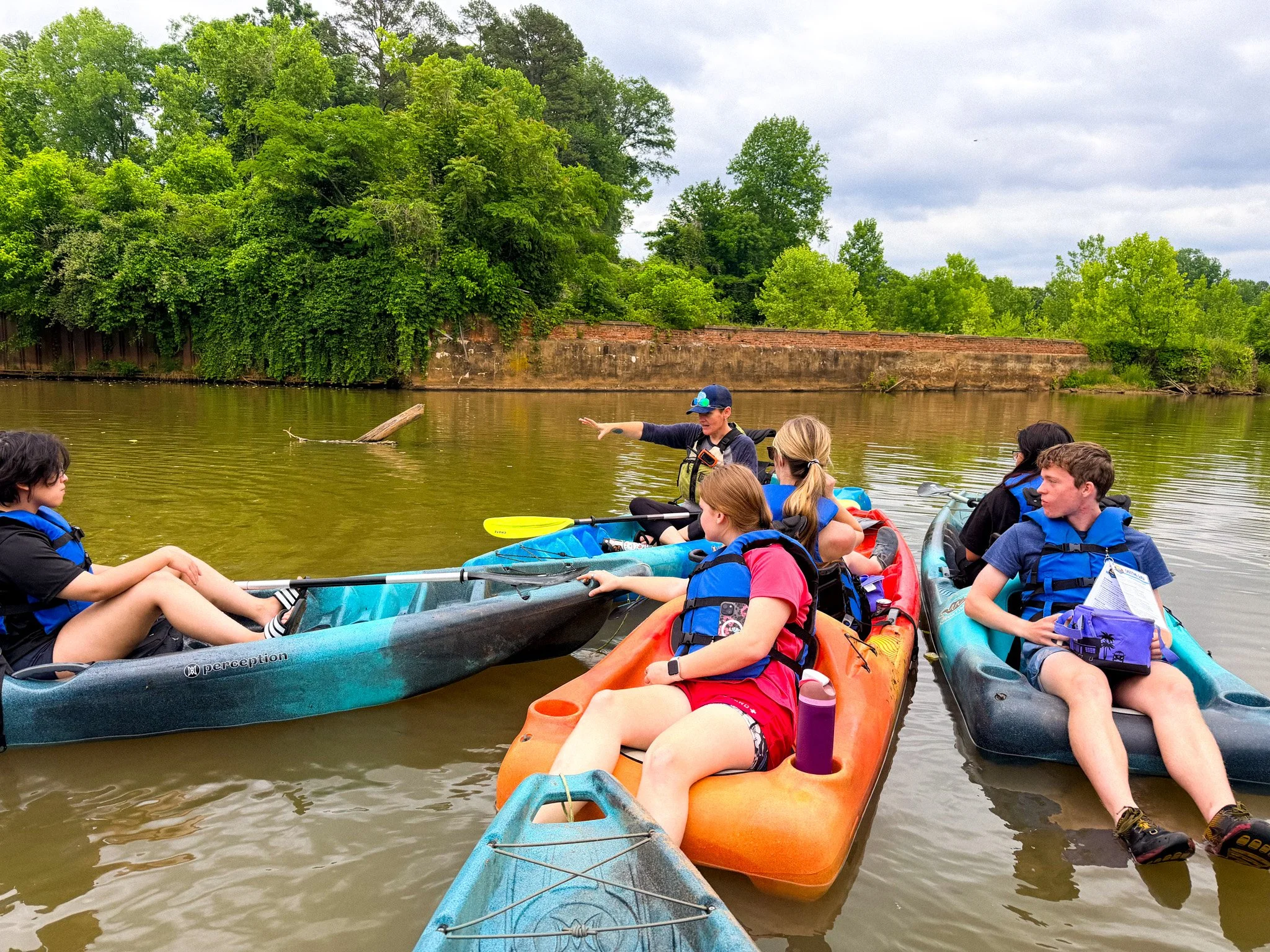 Group of people in kayaks on a river, with a woman standing and giving instructions, surrounded by lush green trees and an overcast sky.