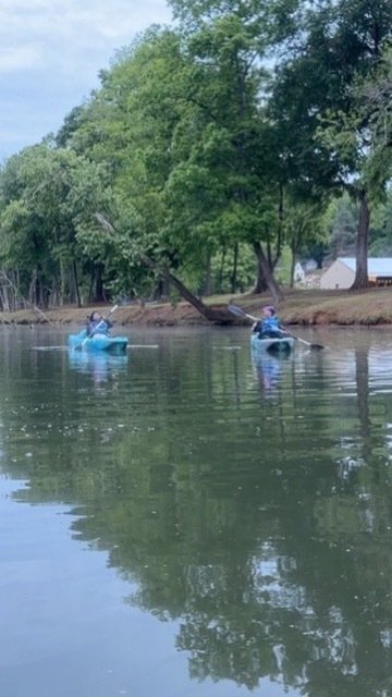 Two people kayaking on a calm river with lush green trees along the bank.