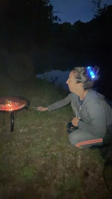 A woman with gray hair styled in a bun, wearing a gray hoodie, is crouching by a bonfire near a body of water at night, holding a stick over the fire.